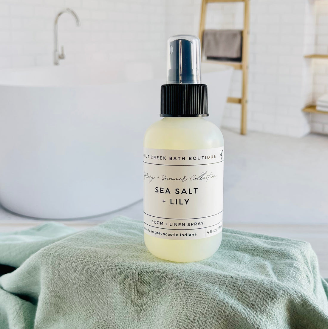 sea salt and lily room and linen spray is in a opaque bottle with a black spray nozzle. it is sitting on a raised platform with a green towel unter it and in the background is a spa like bathtub