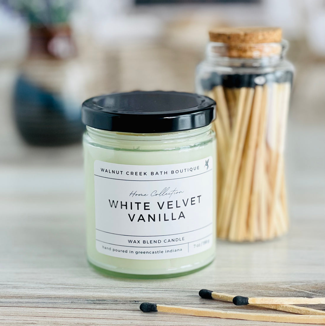 White velvet vanilla candle with black lid. there are a few matches scattered in front and a pretty glass jar of matches in the background