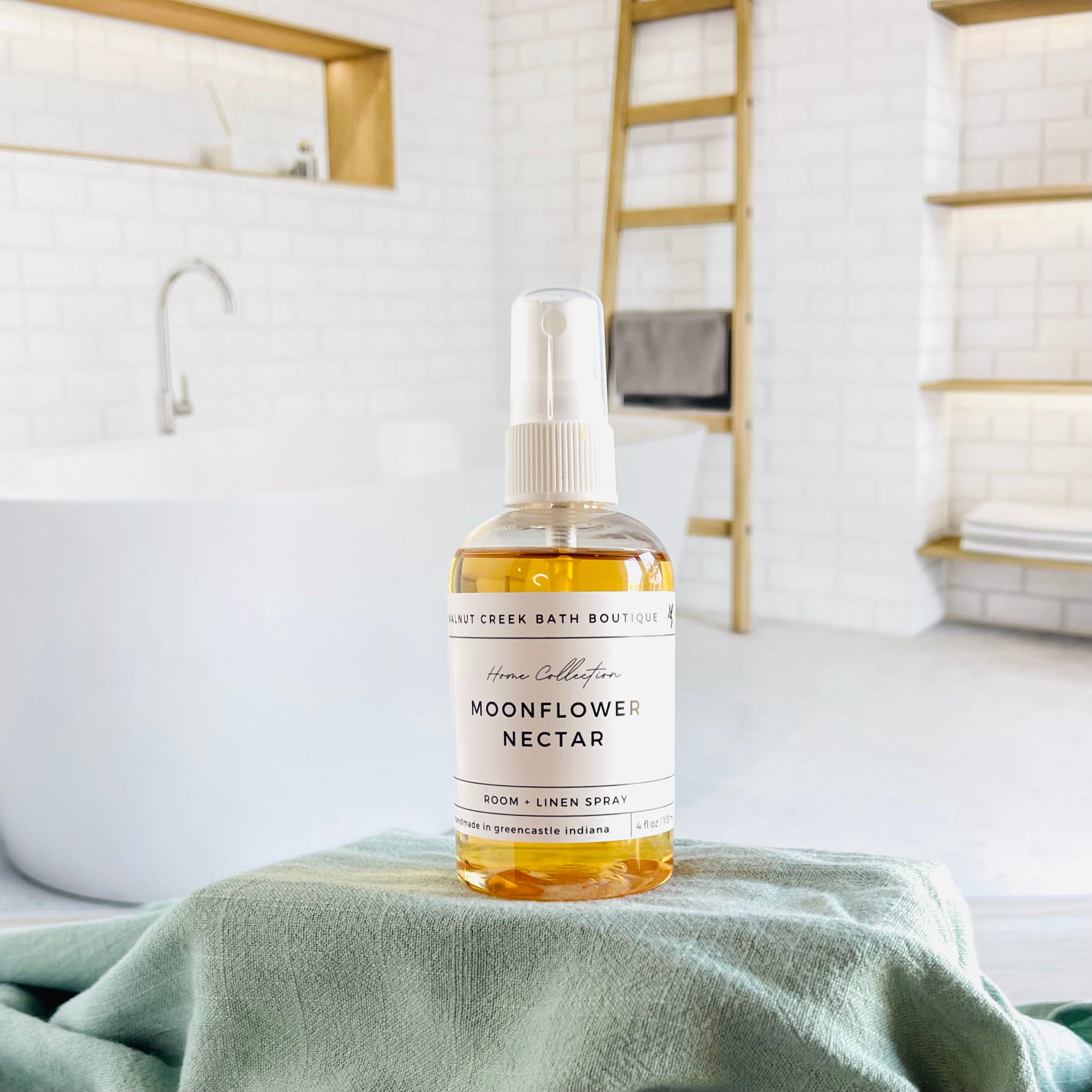 moonflower nectar room and linen spray is in a clear bottle with a white spray nozzle. it is sitting on a raised platform with a green towel unter it and in the background is a spa like bathtub