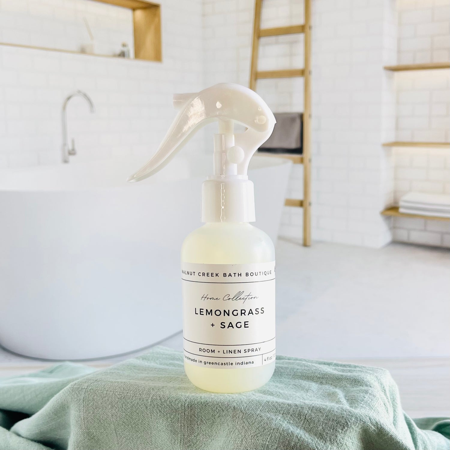 lemongrass and sage room and linen spray is in a opaque bottle with a larger spray nozzle for ease of use. it is sitting on a raised platform with a green towel unter it and in the background is a spa like bathtub