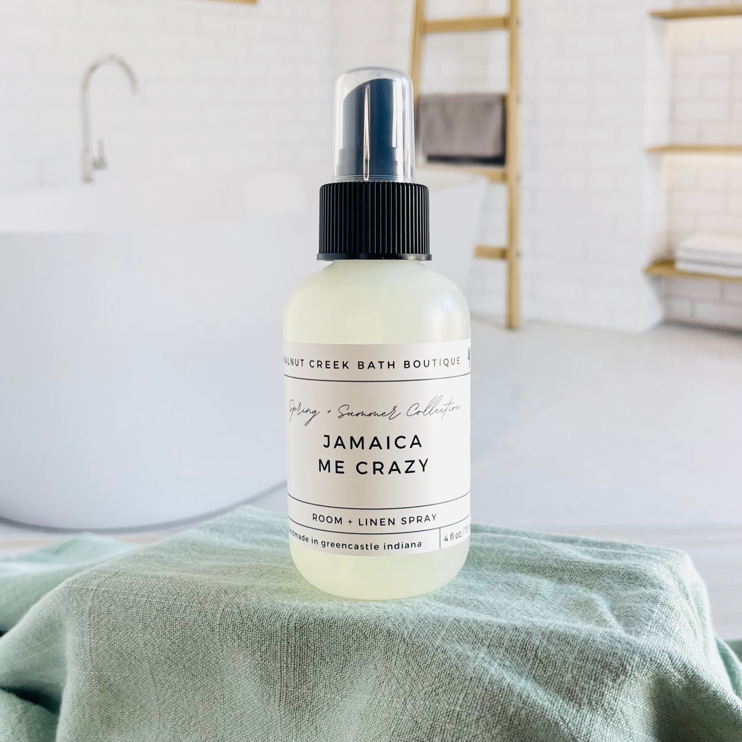 jamaica me crazy room and linen spray is in a opaque bottle with a black spray nozzle. it is sitting on a raised platform with a green towel unter it and in the background is a spa like bathtub