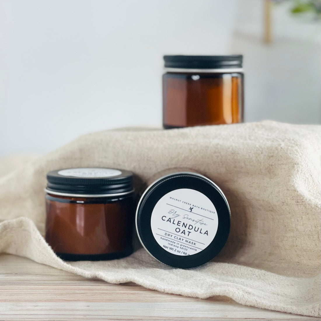 calendula oat face masks are shown, one laying on its side to show the simple label while the other two are standing upright. the mask is in amber glass jars with black lids. 