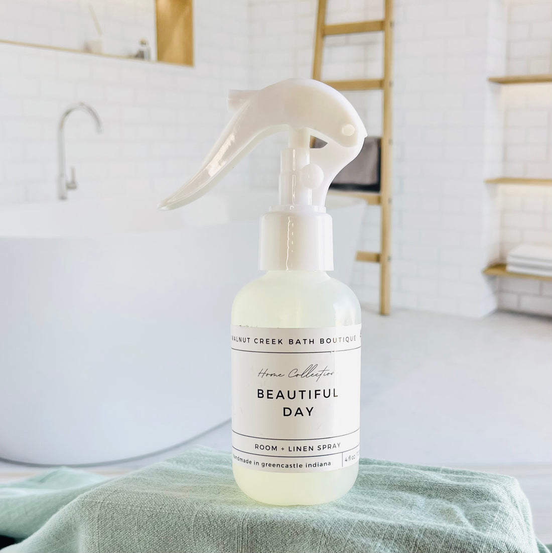 beautiful day room and linen spray is in a opaque bottle with a larger spray nozzle for ease of use. it is sitting on a raised platform with a green towel unter it and in the background is a spa like bathtub