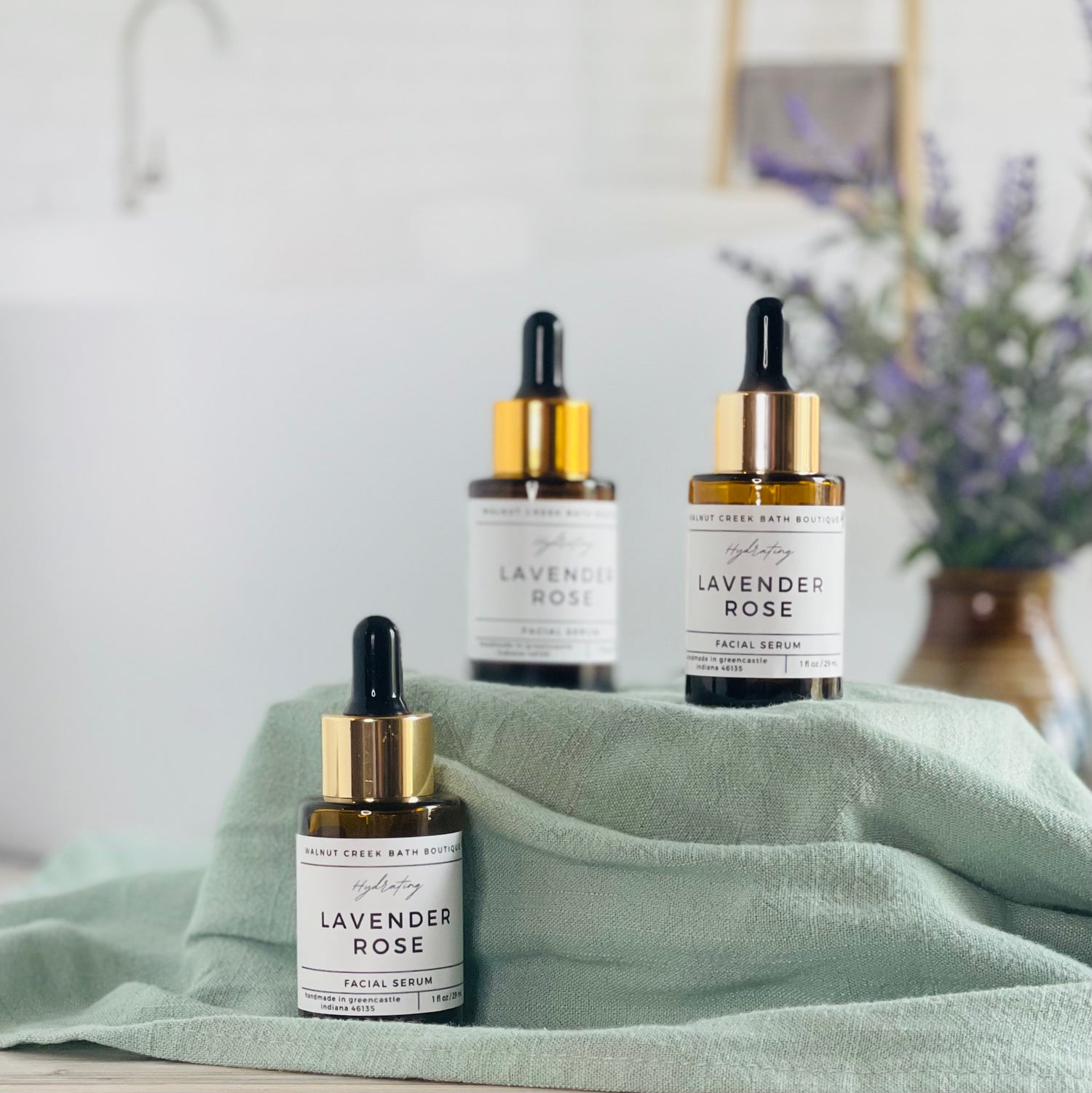 there are 3 lavender rose facial serums shown, one front and center in sharp focus while the other two are lifted on a box with a pretty green towel. these are a little out of focus. in the background is a pretty brown vase with some lavender buds