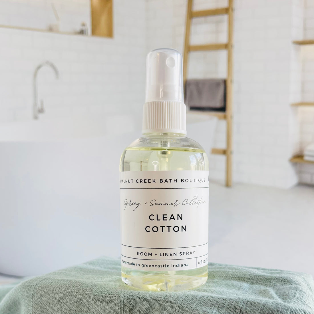 clean cotton room and linen spray is in a clear bottle with a white spray nozzle. it is sitting on a raised platform with a green towel unter it and in the background is a spa like bathtub