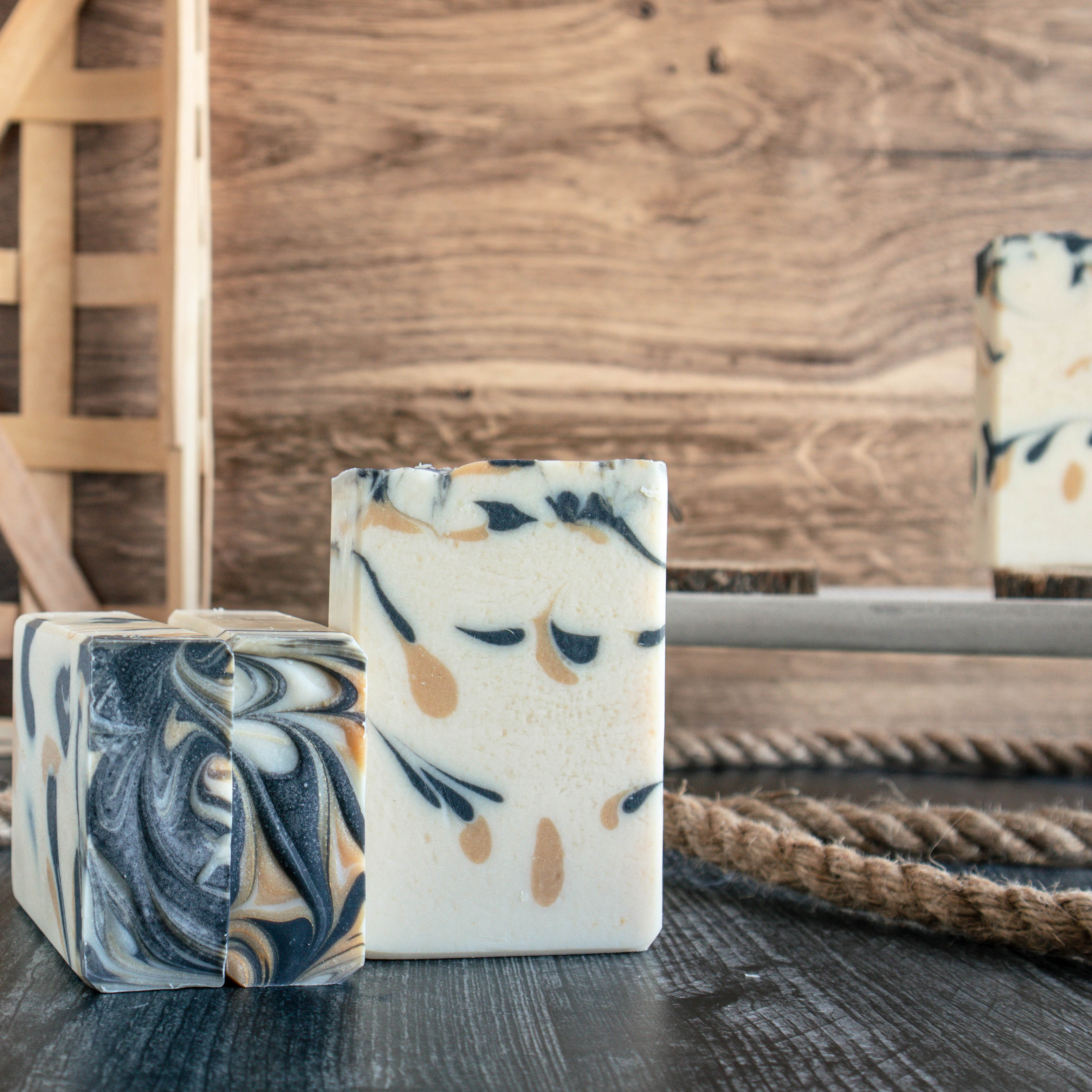 Teal + Cardamom soap showing cream color with drops of gold and black throughout. To the left are 2 bars laying flat showing the pretty black and gold swirls. There is a rope running along behind them with a woven basket in the background on the left.  