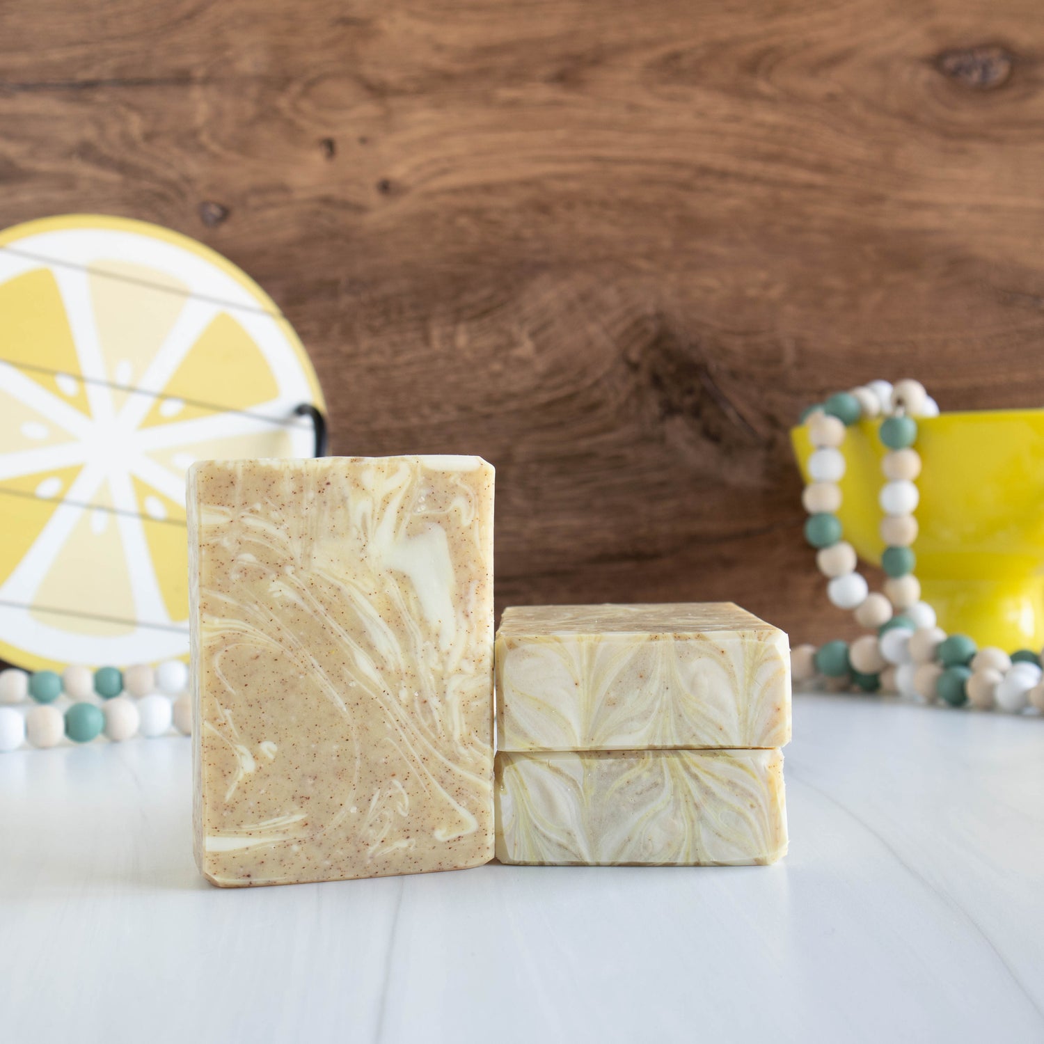 a lemon verbena soap standing tall showing yellow and cream swirls. two more are laying flat to show the pretty tops. there is a yellow Lemon plat in the background and another yellow bowl with some wooden beads coming out of it on the back right.