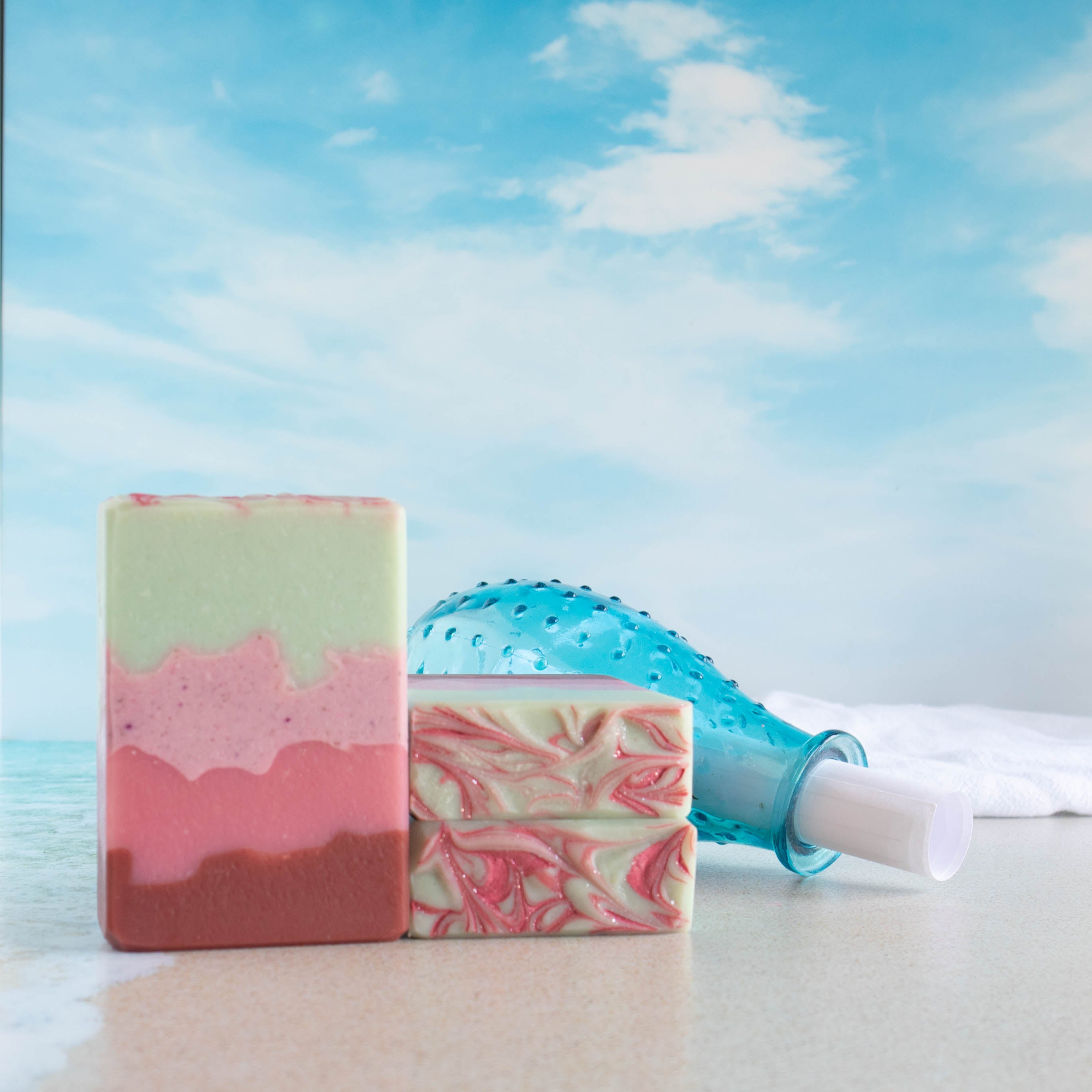 a pink sands soap is standing on a beach scene with waves lapping at its bottom. the soap has 3 pink layers starting dark to light from the bottom. the top layer is a pretty minty green. 2 soaps are laying flat to show a pretty pink swirl on the green top soap. there is a bottle in the background with a letter sticking out of it.