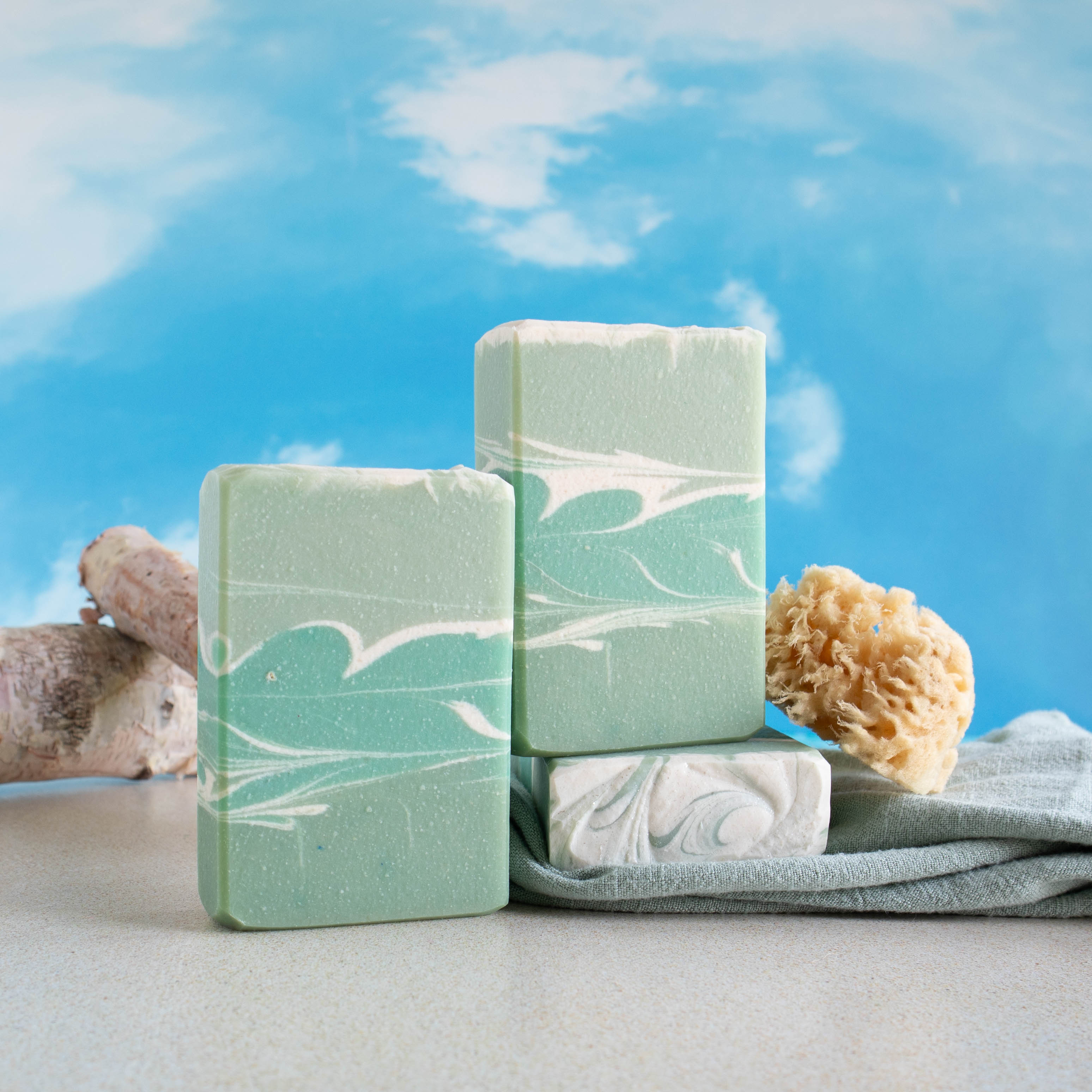 ocean breeze soaps are a light green with a swath of darker green in the middle with a small amount of white swirls throughout. There are 2 bars facing you with one sitting on another that is laying flat showing the pretty top. There is a sky background with some white birch logs in the background and a loofah to the right.
