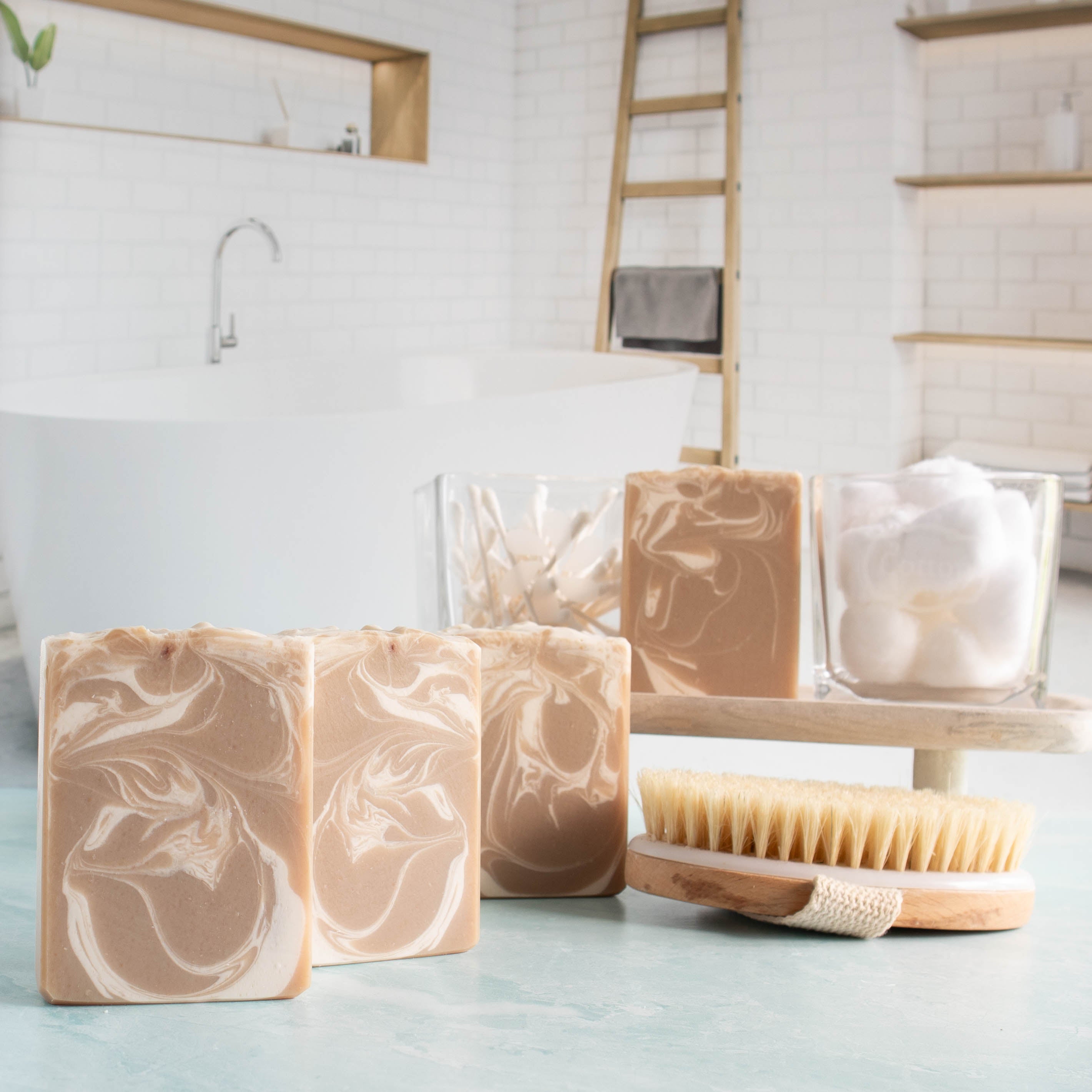 Caribbean teakwood soaps are lined up in a diagonal. they have a very pretty tan color with a wispy white swirl throughout. there is another bar on a tray in the background next to glass containers of q tips and cotton balls. lastly there is a body brush in front of the tray for interest.