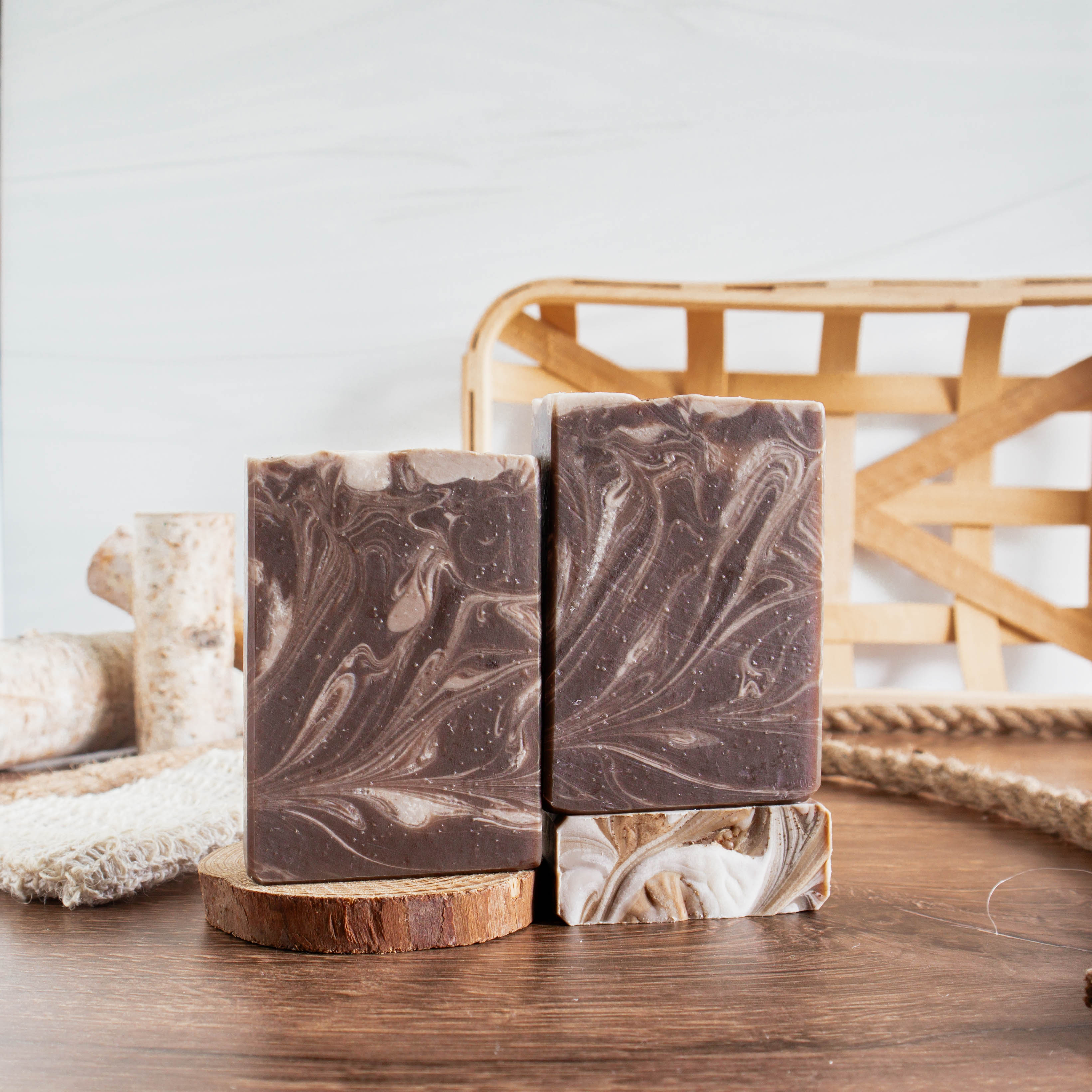 3 bars of Tonka and Oud showing the dark brown soap with different creamy swirls throughout. 2 bars are facing forward and one is laying flat to show the top of the bar. There are small wood logs, rope and a woven basket in the background for interest.