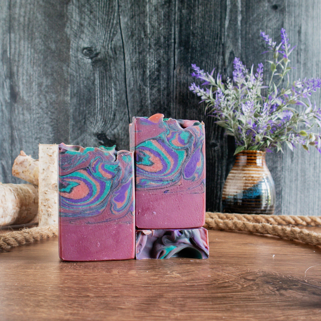 3 bars of black amber and lavender soap showing the reddish bottom third and green, purple and orange swirls on top. One bar is laying flat to show a scoopy top. there is a black rusted wood backdrop and a walnut wood base with some rope, small wood logs and a vase of flowers in the background.