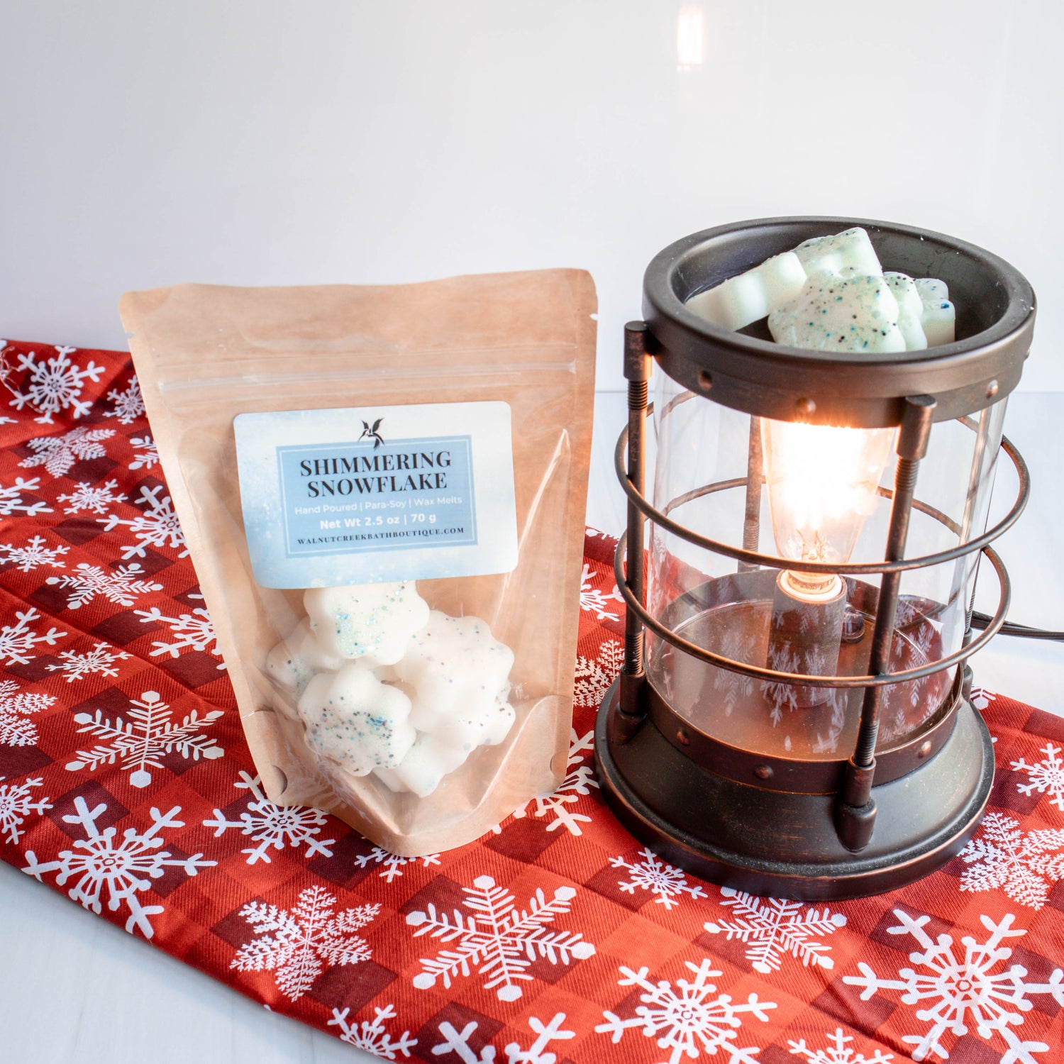 shimmering snowflake wax melts are cute snowflake forms in a bag. the bag is next to a burner with the snowflakes in the warming cup and they are sitting on a red cloth with snowflakes on it. White background