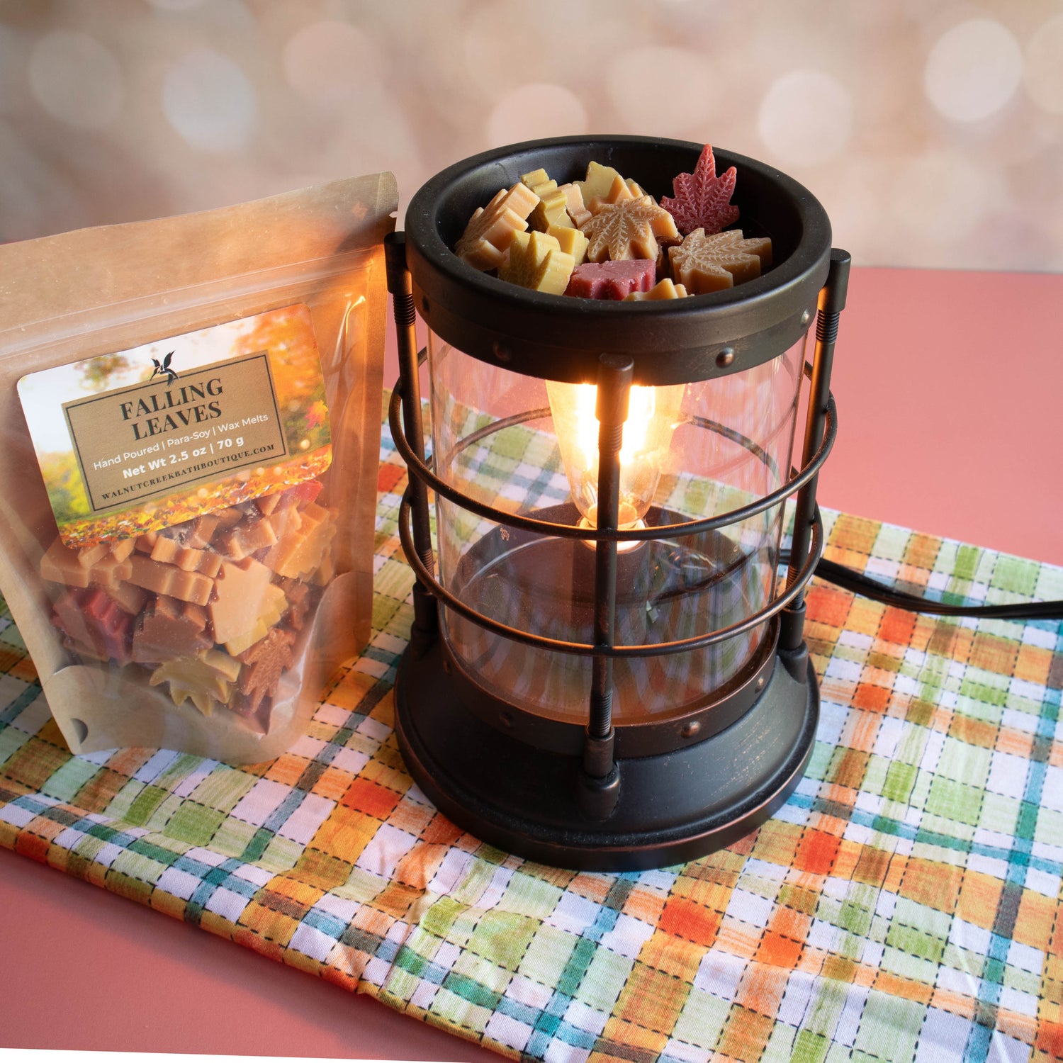 falling leaves wax melts in their bag sitting next to a wax burner filled with the small melts. Colors of red, yellow and orange!. This is sitting on a plaid clothe with the fall colors of green, red, orange, yellow and a touch of blue.