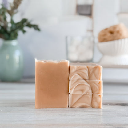 Tan soaps, three stacked and one standing tall on wood base with a vase of flowers in the background