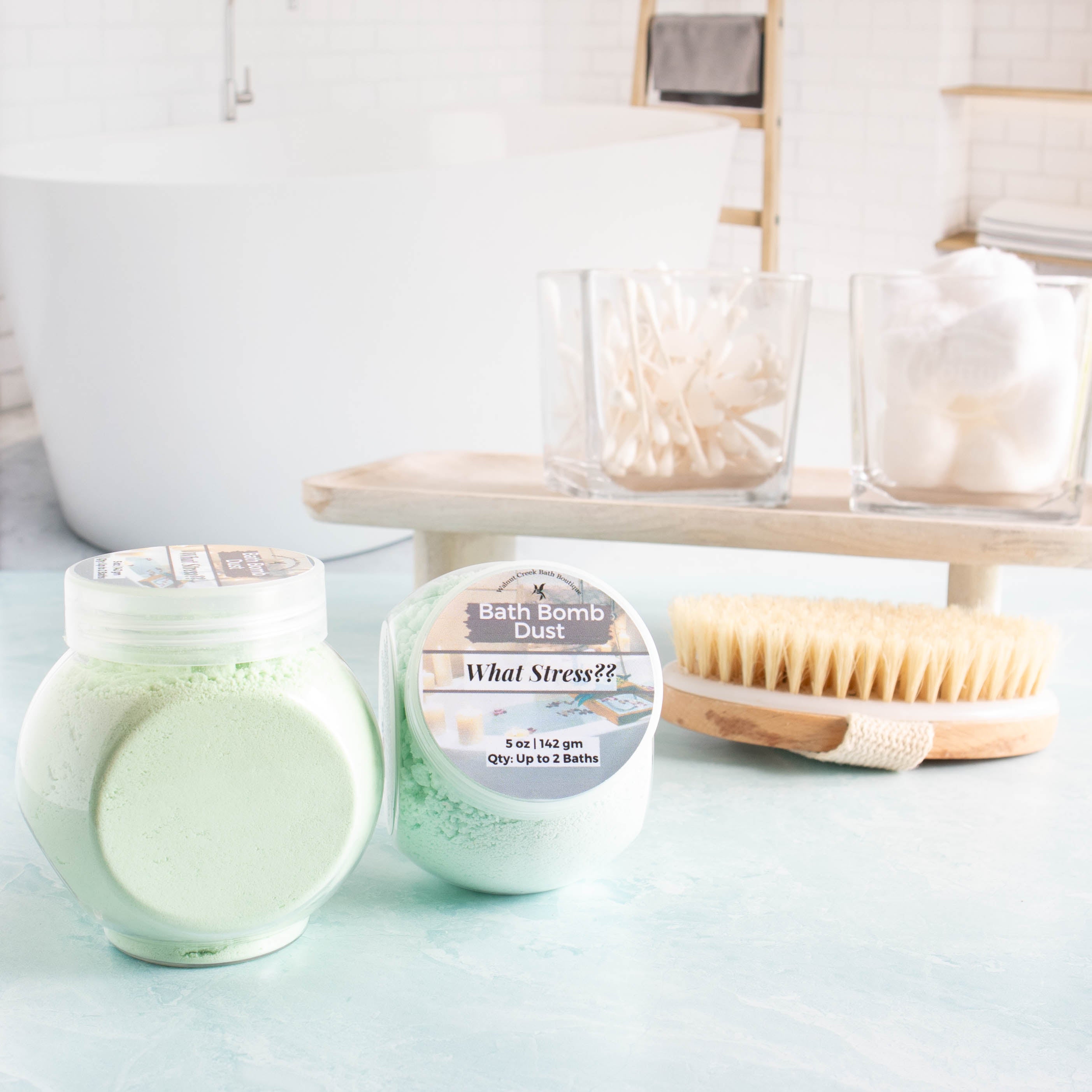 two  5 oz jars of green What stress?? bath bomb powder in the foreground. One showing the label and another showing the side of the jar with the powder.In the background is a tray with cotton balls and q-tips and a body brush. there is a spa like bath room in the background