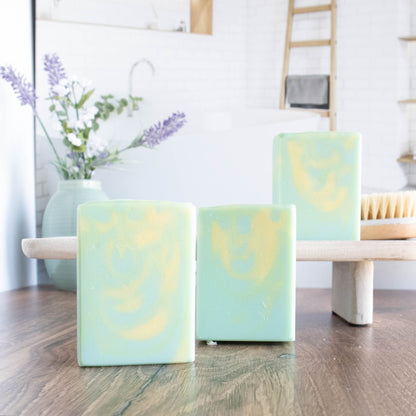 3 agave nectar and lime blossom soaps are shown. they are a light green with a yellow swirl. Ons soap is on a wooden stand. there is lavender flowers in a green vase in the background along with a body brush. there is a bathroom scene in the background as well.