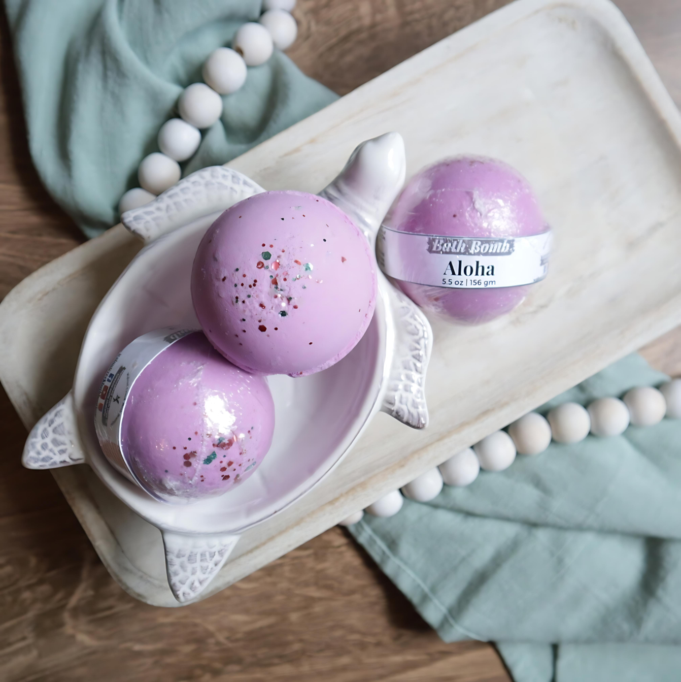 3 aloha bath bombs are shown sitting on a tray. two are in a soap dish in the shape of a turtle. the bath bombs are rosey pink in color with purple pink and green glitter on the tops. there is a warm wood base and a green towel with white beads running under the tray and through the picture.