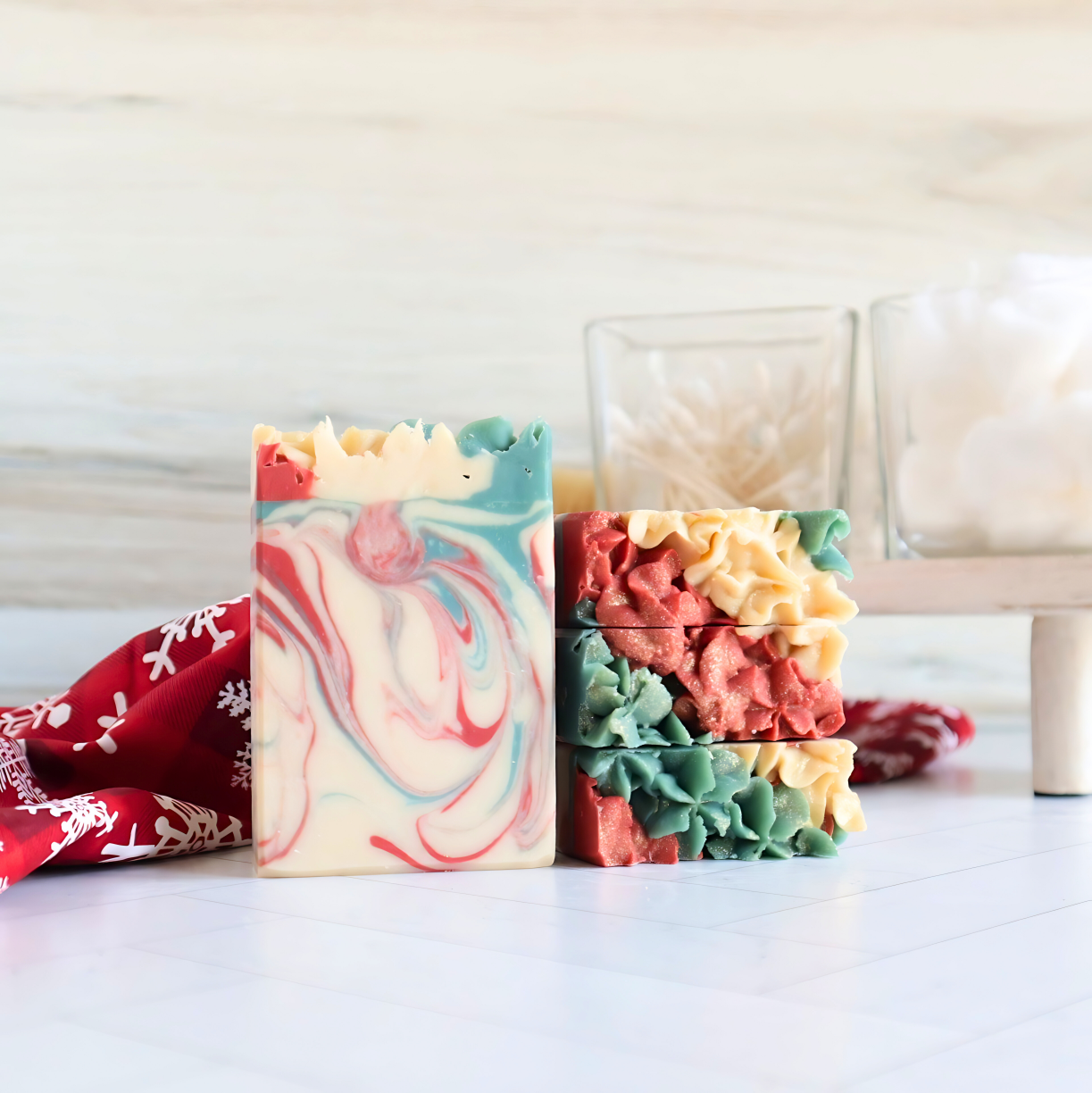 alpine cheer soap is shown, one standing facing front showing the white base soap with pretty red and green swirls. three more soaps are laying flat to show the pretty red, green and white piping on the tops of the soaps. there is a red cloth behind the soaps that has a snowflake design. in the background are a couple of glass dishes on a tray holding q-tips and cotton balls 