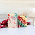 alpine cheer soap is shown, one standing facing front showing the white base soap with pretty red and green swirls. three more soaps are laying flat to show the pretty red, green and white piping on the tops of the soaps. there is a red cloth behind the soaps that has a snowflake design. in the background are a couple of glass dishes on a tray holding q-tips and cotton balls 