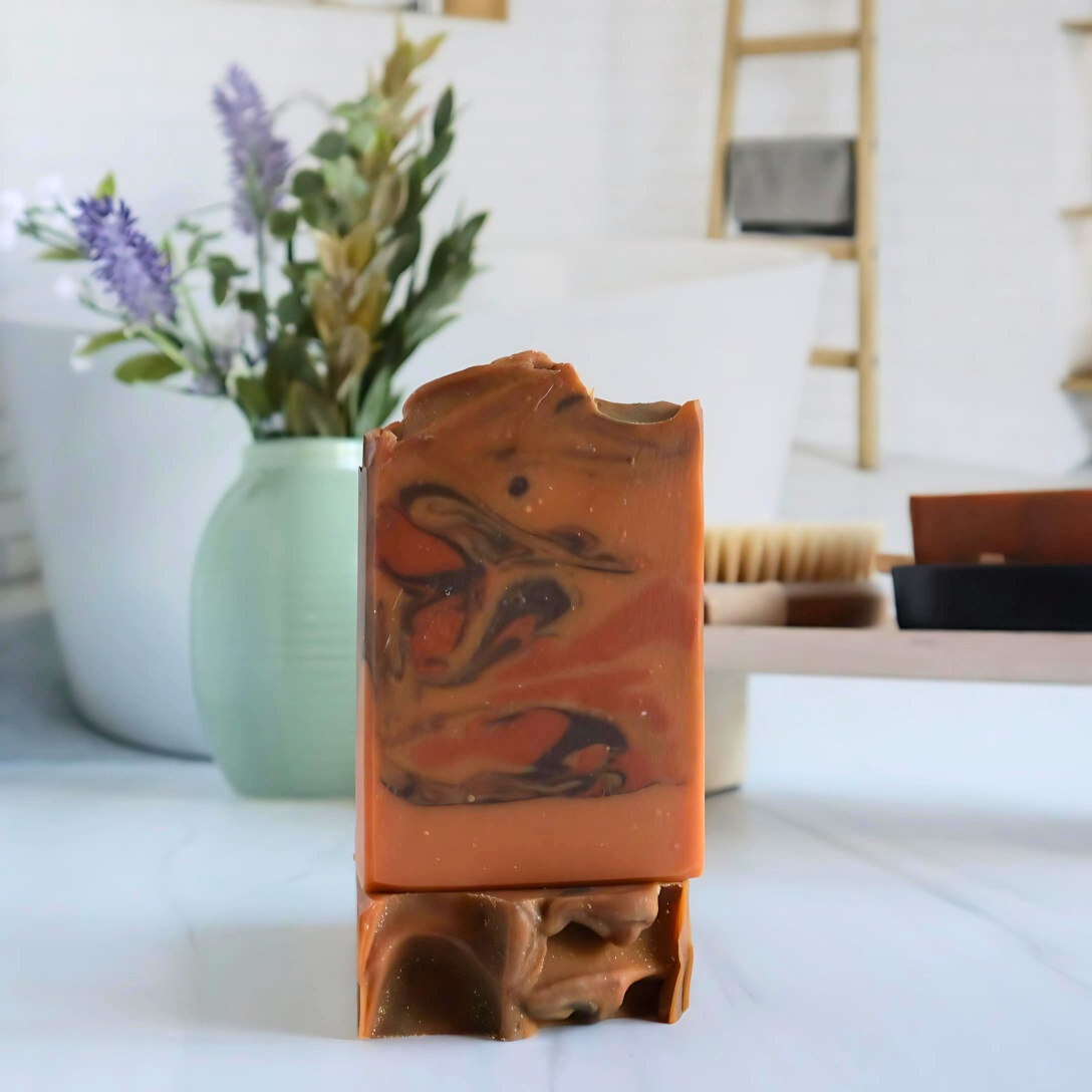 2 ancient incense soaps are shown, one standing tall to show the rusty orange base color with dark brown and dark orange swirls. this is standing on another soap with the top facing you to show the textured top. in the background is a green vase, and a tray with a soap dish.