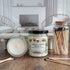 2 jars of apple cider donut candles are shown, one with the lid off and laying flat to show the smooth top and the other with the lid on and label facing you to show that. there are matches in a clear jar to the right of the candles and a few matches scattered around the table as well. in the background is a fireplace flanked by 2 wicker chairs