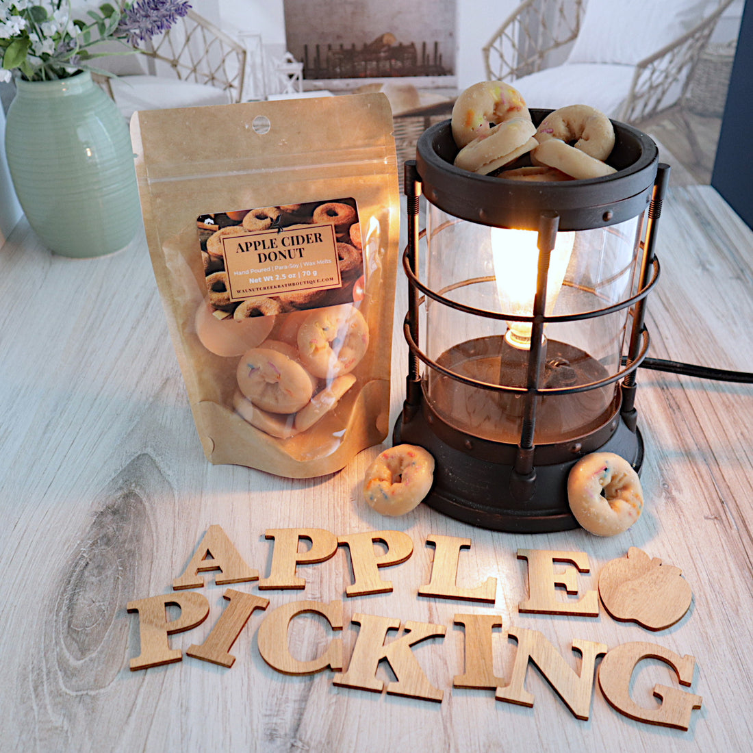 apple cider donut wax melts in a bag is standing next to a lit wax melter with the donut wax melt shapes in the melter cup. there are a couple of donut wax melts on the bottom of the burner. in front of the bag and the burner are wooden letter spelling out &quot;apple picking&quot;.  this is all sitting on a washed out wooden base with a couple of chairs in the background next to a fireplace
