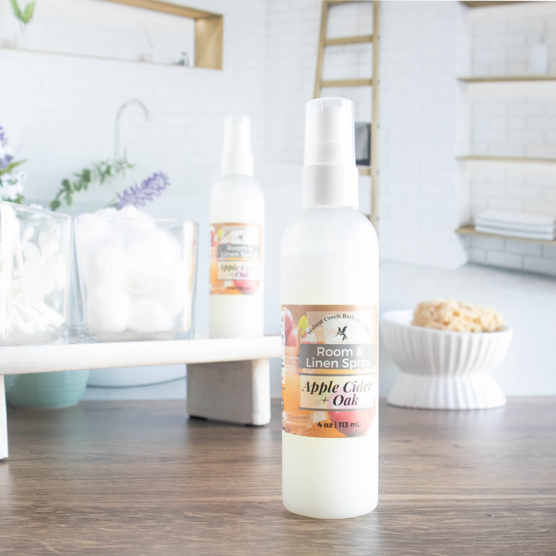 apple cider and oak room spray is front and center. it is in a squat round bottle with black sprayer. in the background is a tray with some cotton balls and q-tips in jars along with another room spray. in the background is an image of a spa bathroom and a dish with a loofah