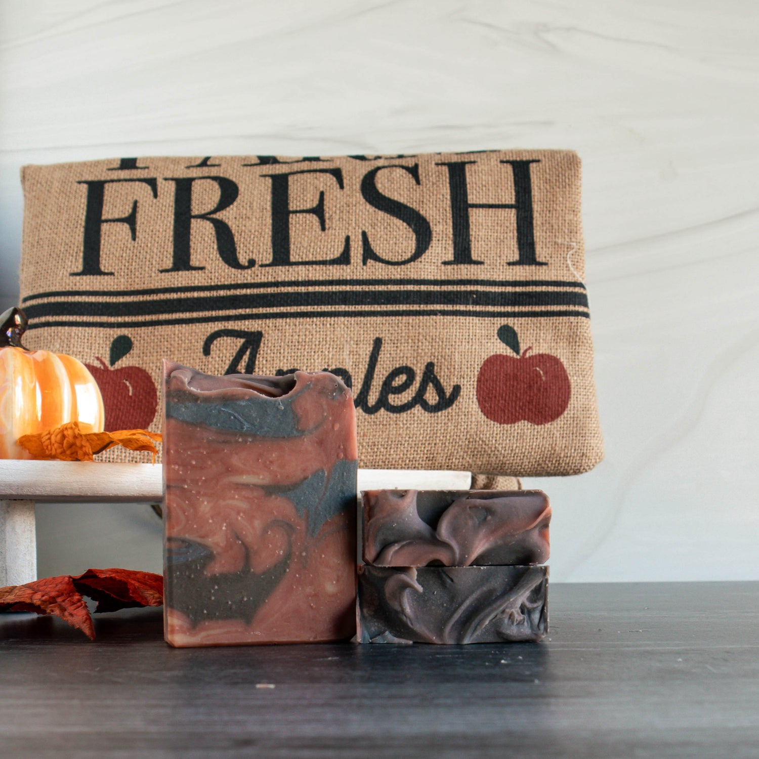 one apple cider oak goat milk soap is standing next to 2 others laying flat. The soap is a dark fire red with swirls of dark blue and black. there is a burlap apple sack in the background along with some pumpkins and leaves