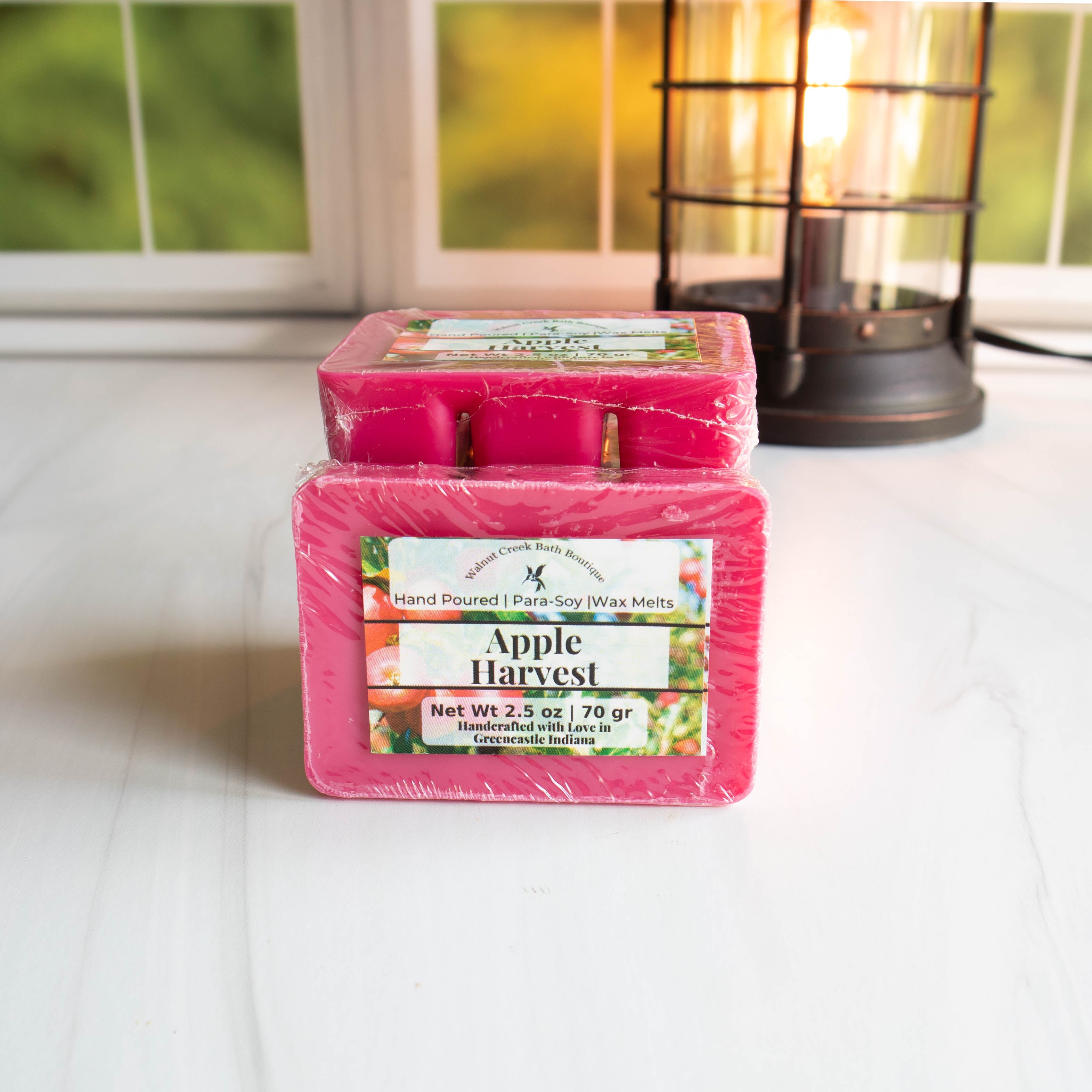 Apple harvest wax melts stacked with one facing you. They are red n color and sitting on a white base in front of a window with a wax burner in the background all lit up.