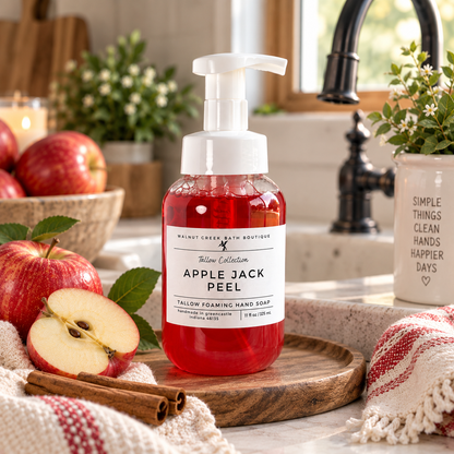 A bottle of Apple Jack Peel foaming hand soap by Walnut Creek Bath Boutique, filled with red liquid and topped with a white pump, set on a wooden board in a kitchen scene with apples, cinnamon sticks, and a towel.