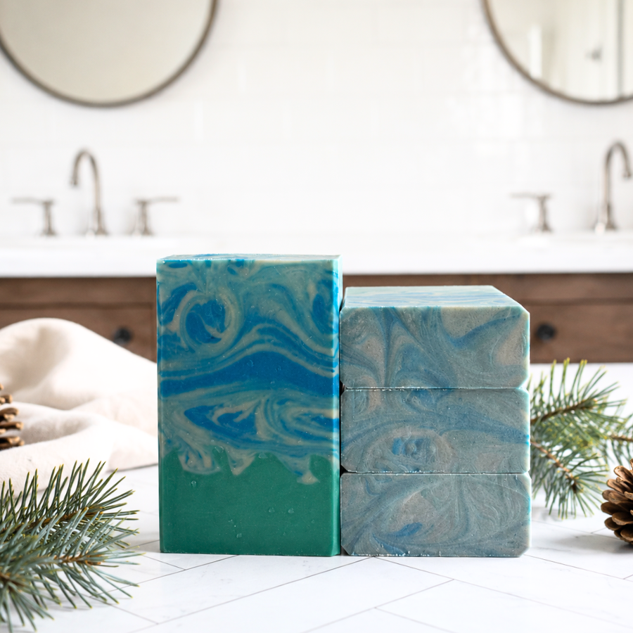 Two handmade marbled soaps, one tall and one stacked, in shades of teal, blue, and white, styled on a white tiled countertop with pine branches and a soft cloth in a bathroom setting.