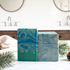 Two handmade marbled soaps, one tall and one stacked, in shades of teal, blue, and white, styled on a white tiled countertop with pine branches and a soft cloth in a bathroom setting.