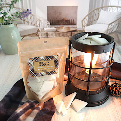 autumn flannel wax melt bag is standing to the left of a lit wax melt burner. there are wax pieces in the burner. there are also wax pieces piled next to the burner and bag.  there is an autumn colored fabric running along under the bag and behind the burner. this is all sitting on a washed out wooden base with a couple of chairs in the background next to a fireplace