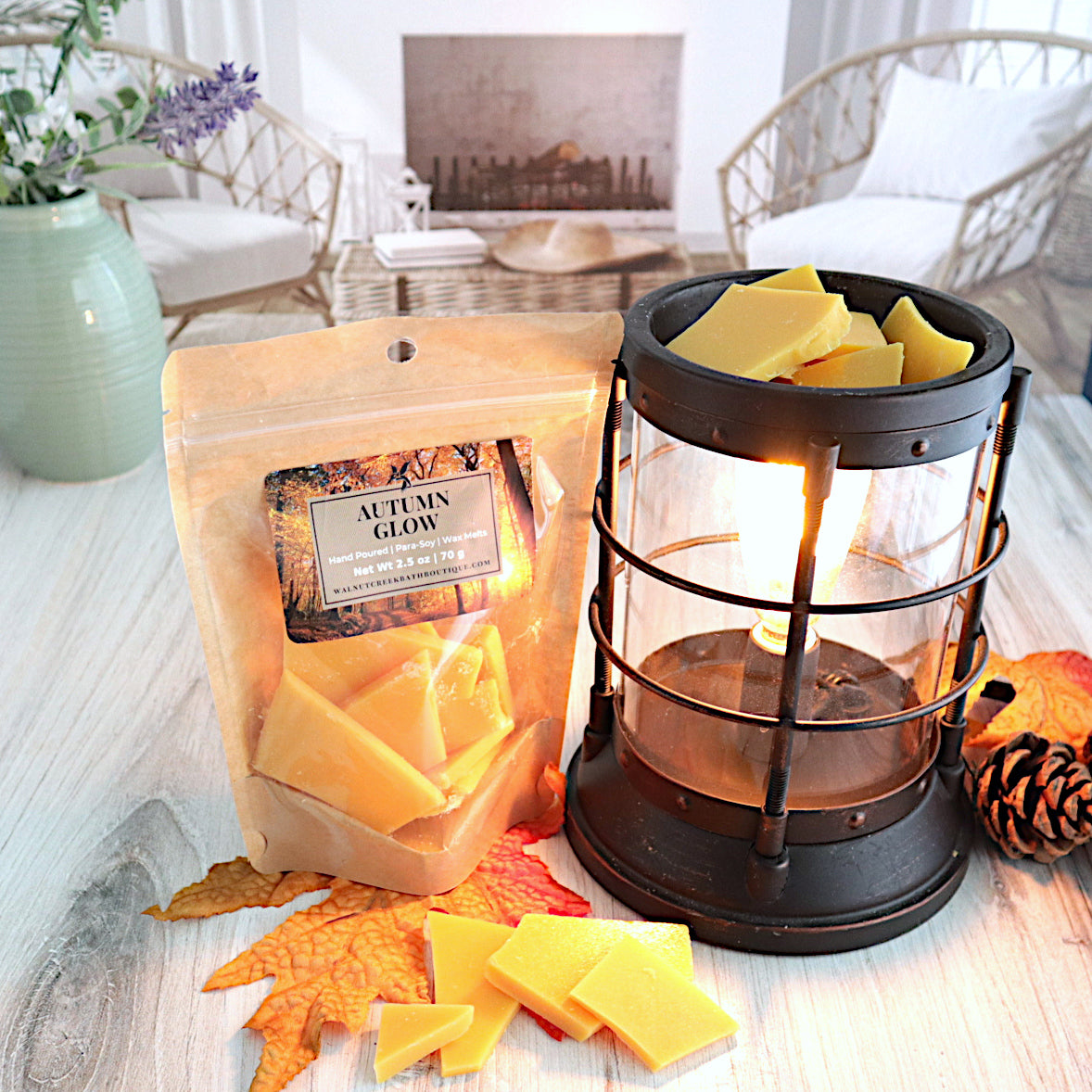 autumn glow wax melt bag is standing next to  a wax burner with wax pieces in it waiting to melt.  the wax is orange pieces of small to medium size. there is a fall leaf on the the surface that the bag and pieces are covering some of it. there is also a pinecone on the right side of the burner.  this is all sitting on a washed out wooden base with a couple of chairs in the background next to a fireplace