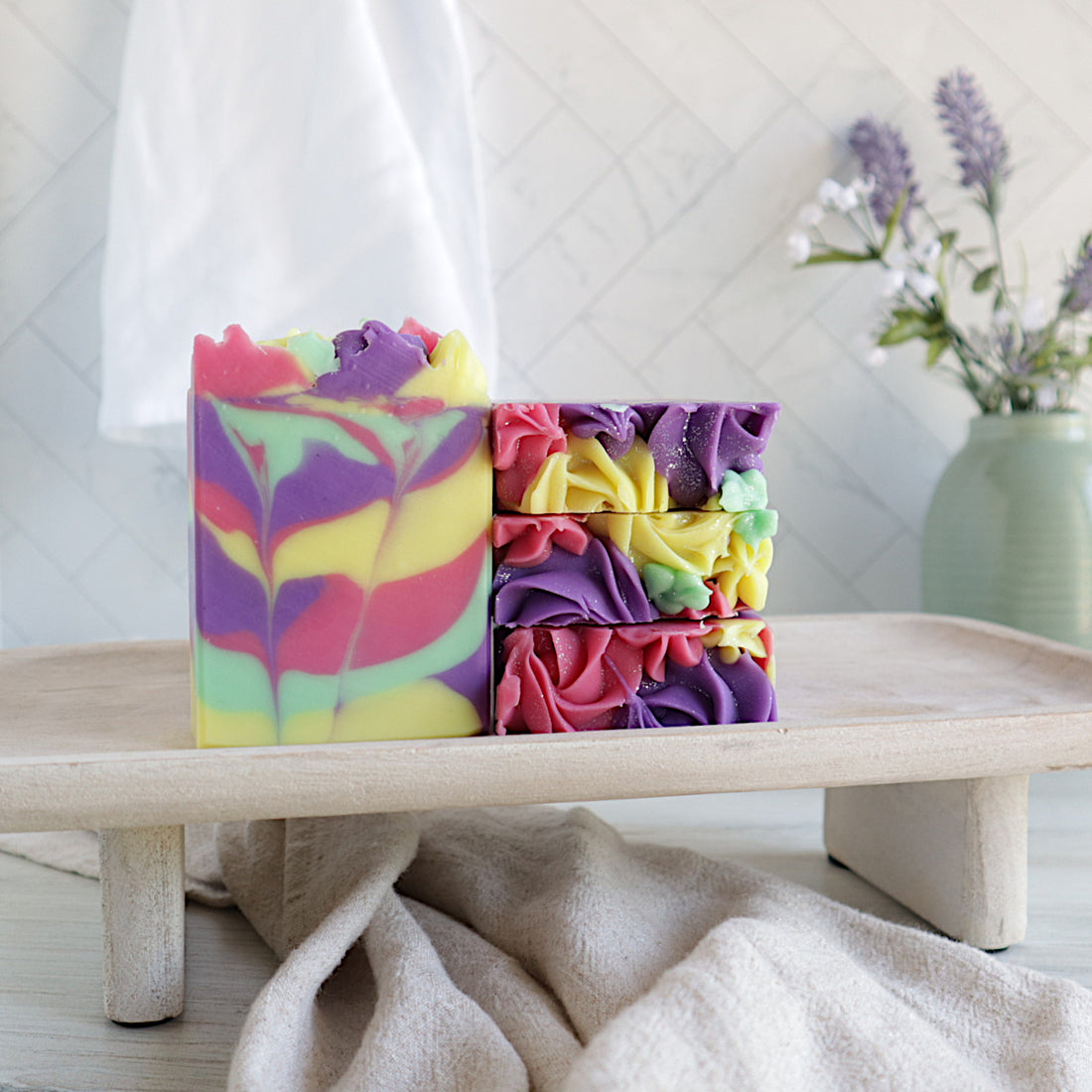 there is 1 soap facing forward showing the pretty swirls in colors of yellow, purple, green and pink. next to that are 3 more soaps stacked on top of each other showing the piped tops of all the colors. they are all on a riser with a towel running unerneath. there is a green vase in the back right with some lavender buds. there is also a white towel hanging in the background