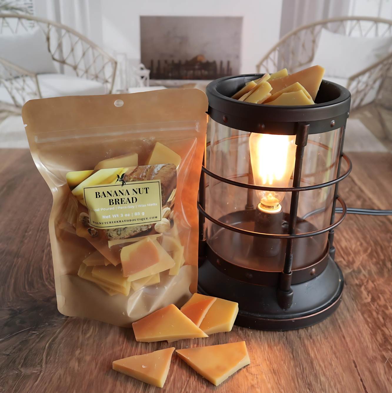 A banana nut bread wax melt bag is standing next to a lit bronze wax melt burner. this has the tan and brown bits of wax in it and there are some more of these scattered along the base. in the background is a fireplace flanked by 2 wicker chairs.