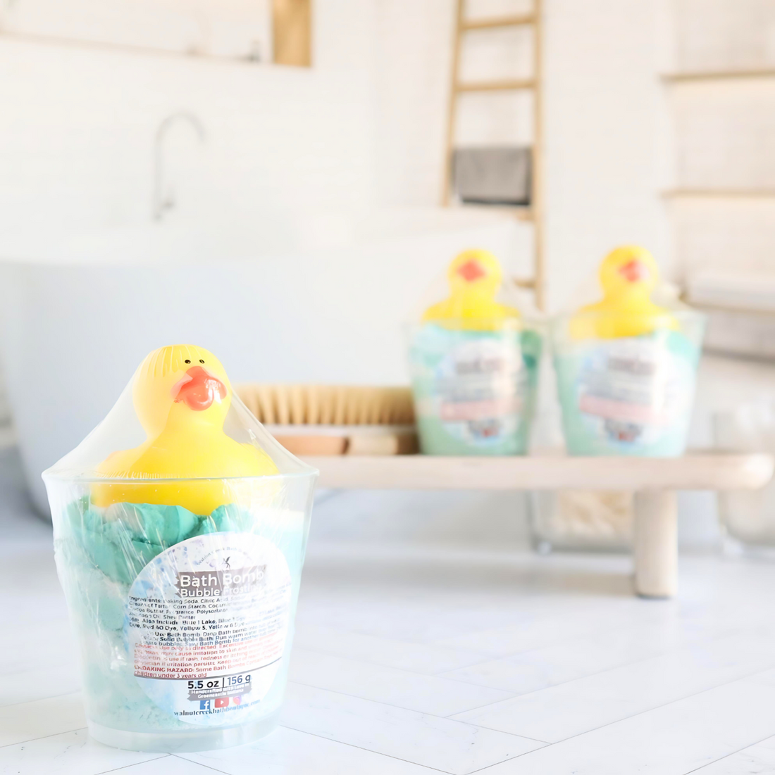 cup with bath green and white bomb powder is toward front, this cup has a cute little rubber ducky on top of some bath bomb frosting. there are 2 more cups in the background on a tray along with a shower brush.