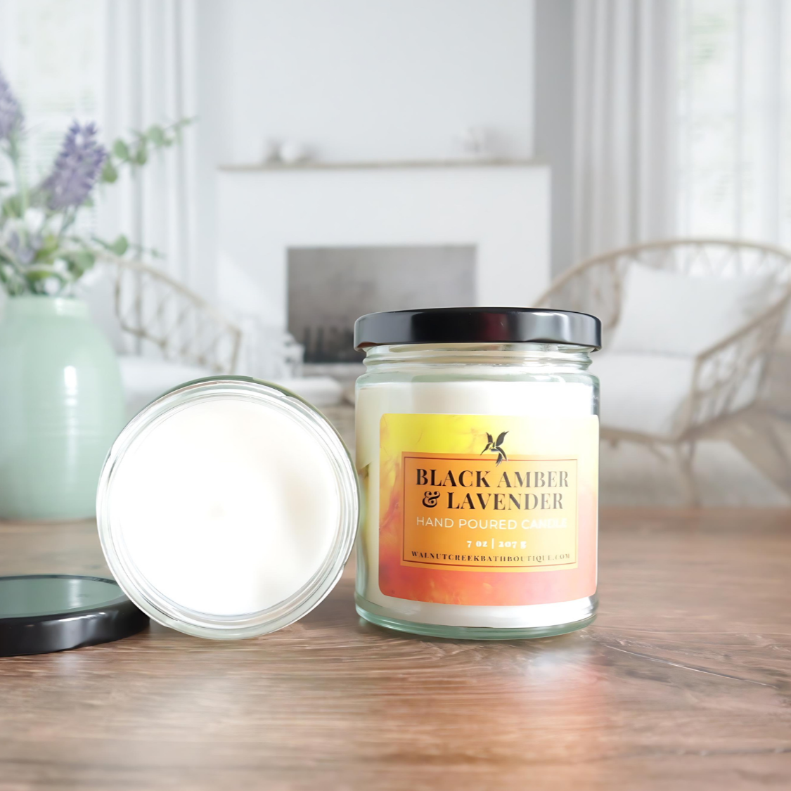 black amber and lavender candle is shown with one open laying flat with the top showing, the lid is off to the side. the other candle is standing next to that one with the lid on and showing the label that is amber in color, with some yellow and orange. there is an image of a fireplace in the background surrounded by 2 wicker chairs. there is also a sage green vase with lavender buds in the back left of the image.