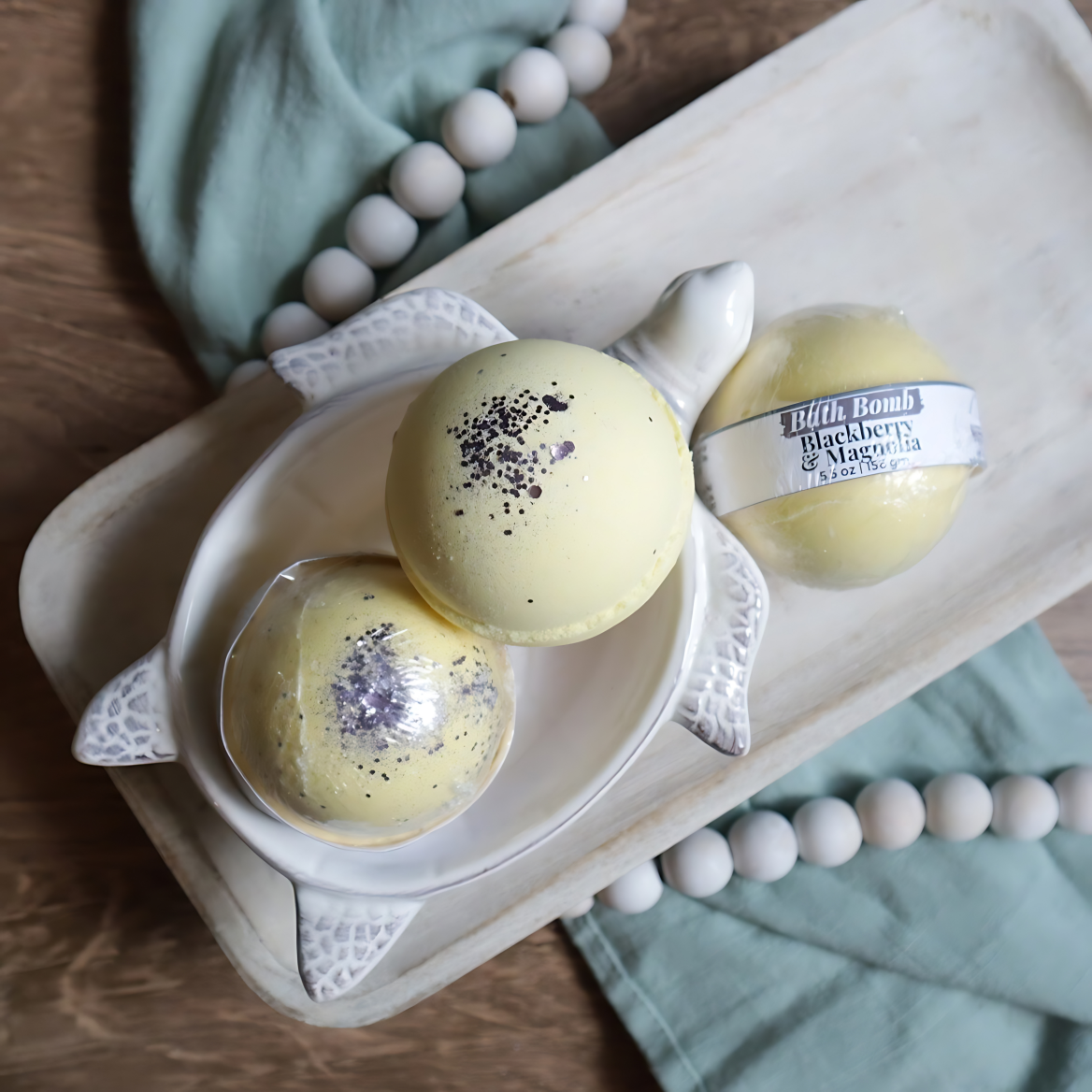 3 blackberry and magnolia bath bombs are shown sitting on a tray. two are in a soap dish in the shape of a turtle. the bath bombs are yellow in color with purple glitter on the tops. there is a warm wood base and a green towel with white beads running under the tray and through the image