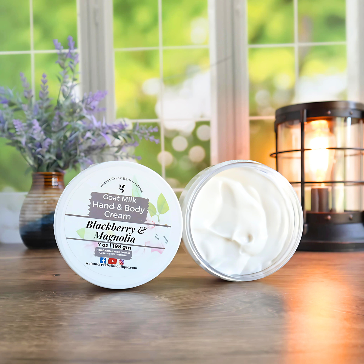 2 blackberry and magnolia creams are shown standing on their sides. One to show the label and the other to show the creamy swirly contents. in the back left is a vase with lavender buds and back right is a lit wax burner to add some light. behind all of this is a window with some greenery showing through.