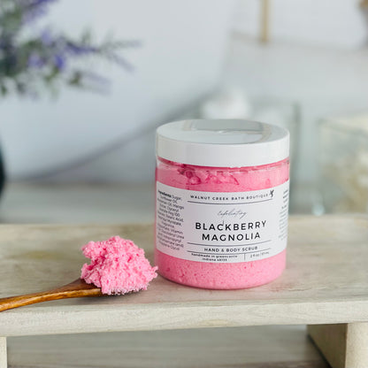 blackberry magnolia scrub is shown on a tray with a wooden spoon mounded with the luscious scrub to show this fluffy consistency.. there is some lavender in the background to give interest to this very calm and neutral image to evoke a peaceful experience. 