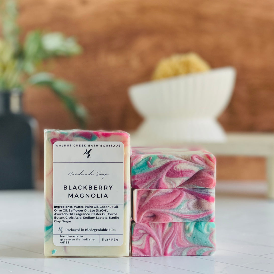 blackberry magnolia soaps are shown, one standing facing out to show the label. Next to this are three more soaps stacked on top to show the pretty swirls. in the background is a soap dish on a stand and a small vase with some greenery.