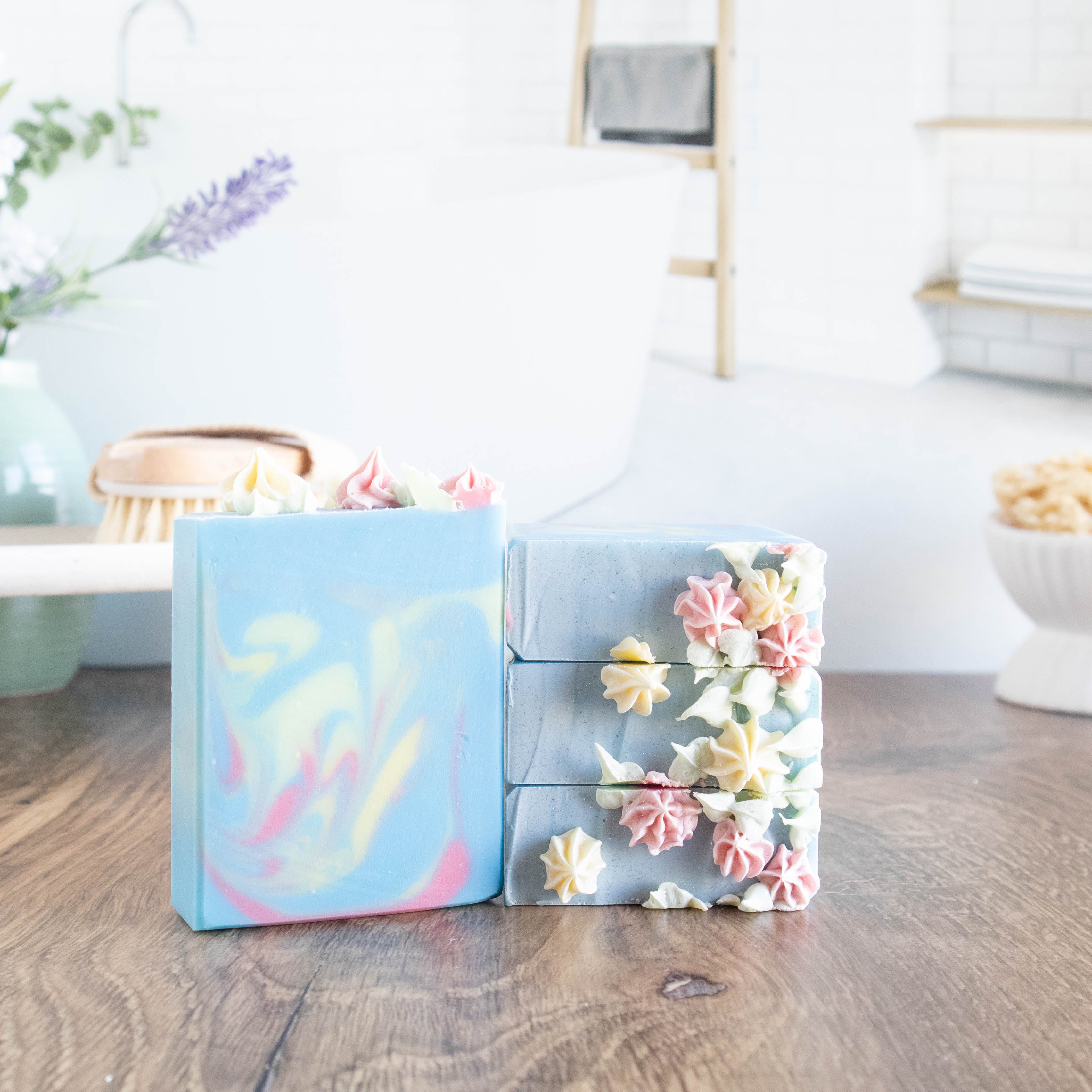 there are three blackberry and magnolia soaps stacked to show the piping. there are small little flowers piped on the top. there is another soap standing to the left of these to show the swirls. he bars are a light blue and the swirls are a soft yellow and pink. in the background on left is a body brush sitting on a wooden tray and on the back right is a white dish with a loofah. in the background is a scene of a lovely bathroom