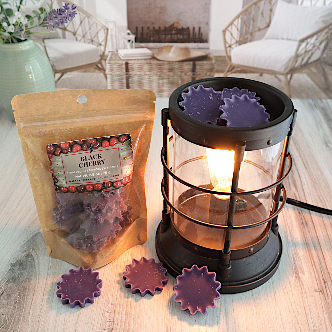 Black cherry wax melts in a bag is standing next to a burner that is lit and also have the cute starburst shaped purple wax melts in the dish just waiting to melt. there are more wax melts on the table in front of the bag and burner. this is all sitting on a washed out wooden base with a couple of chairs in the background next to a fireplace