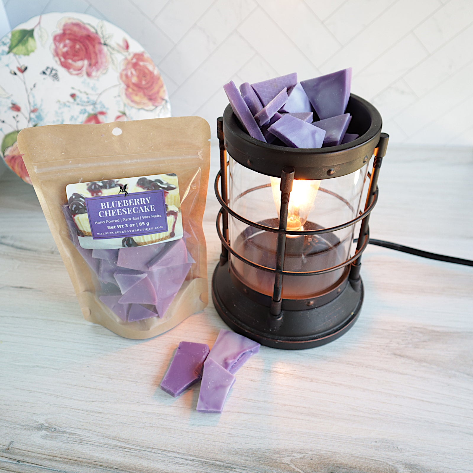 blue berry cheesecake wax melt bag standing next to a bronze burner that is turned on. there are purple shards of wax pieces both in the burner and along the base. there is a pretty floral plate in the background