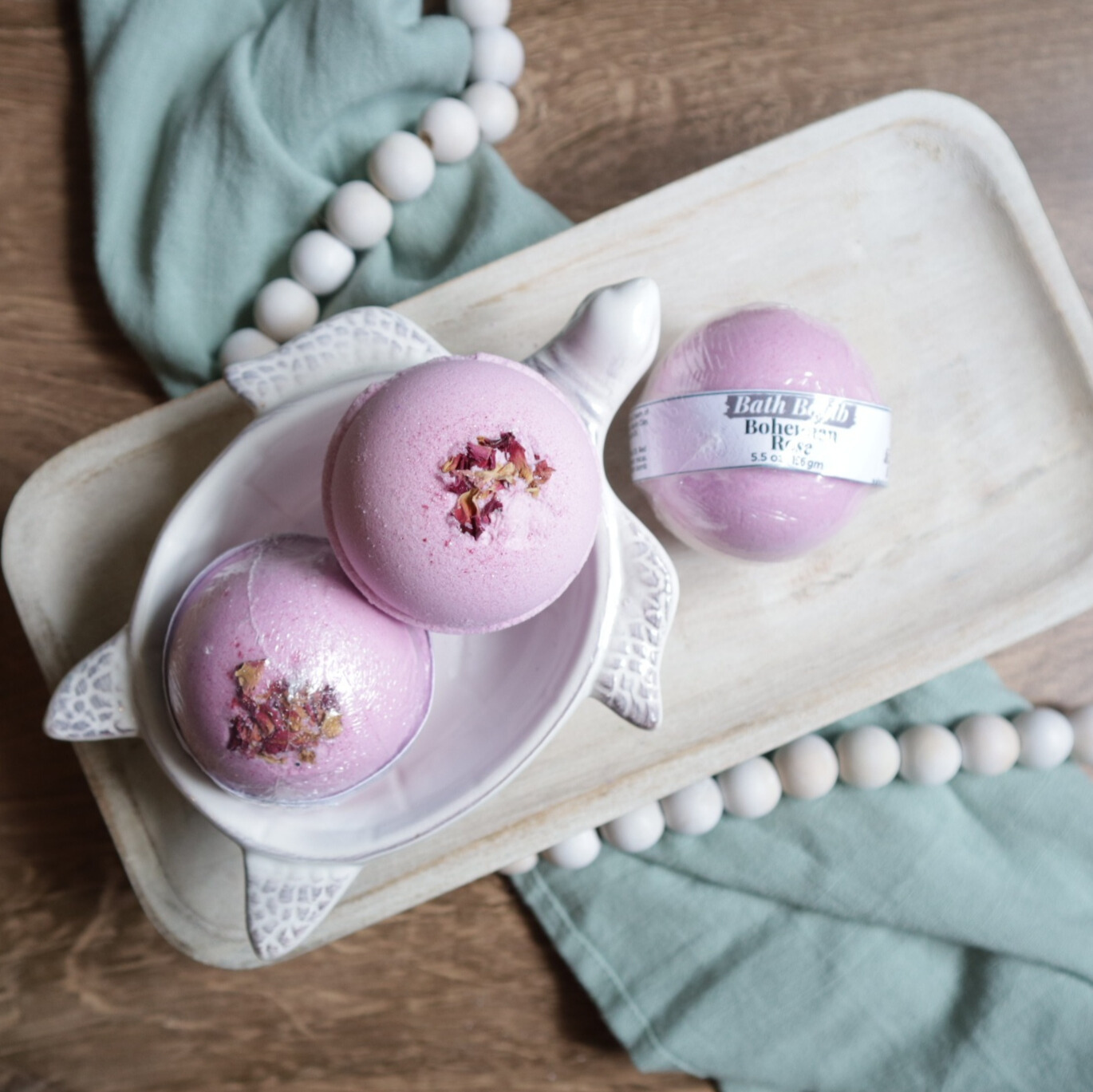 3 bohemian rose bath bombs are shown sitting on a tray. two are in a soap dish in the shape of a turtle. the bath bombs are pink in color with rose buds on the tops along with some pink glitter.  there is a warm wood base and a green towel with white beads running under the tray and through the picture.