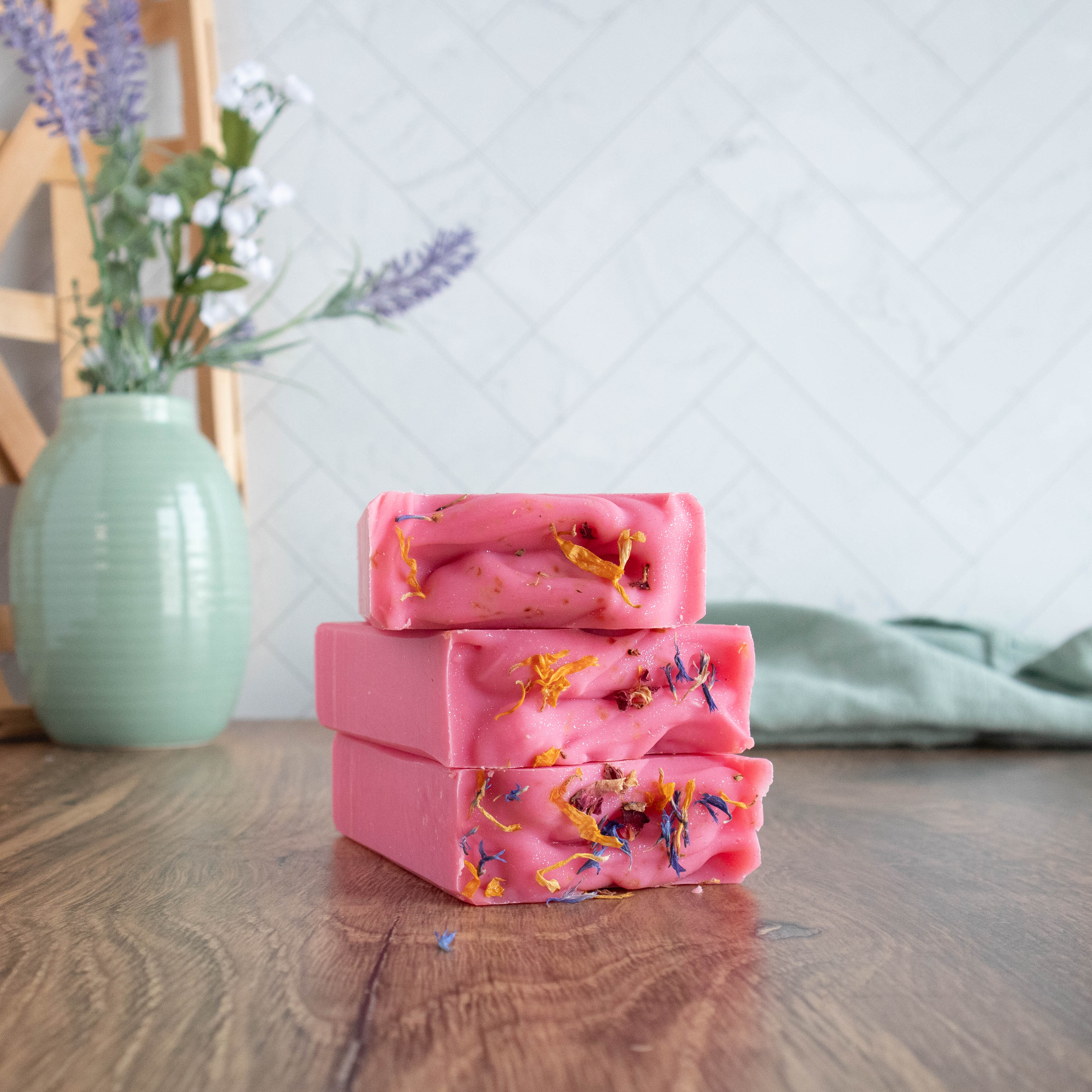 3 very pretty pink soaps stacked on top of each other with the tops facing, showing the sprinkling of blue and yellow flower petals that are adorning the tops. there is a green vase in the background and a green towel running in the back as well