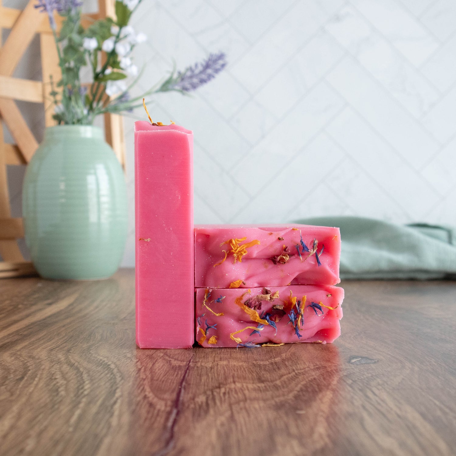 a very pretty pink soap - one is standing showing the side while next to that are two others with the tops facing, showing the sprinkling of blue and yellow flower petals that are adorning the tops. there is a green vase in the background and a green towel running in the back as well