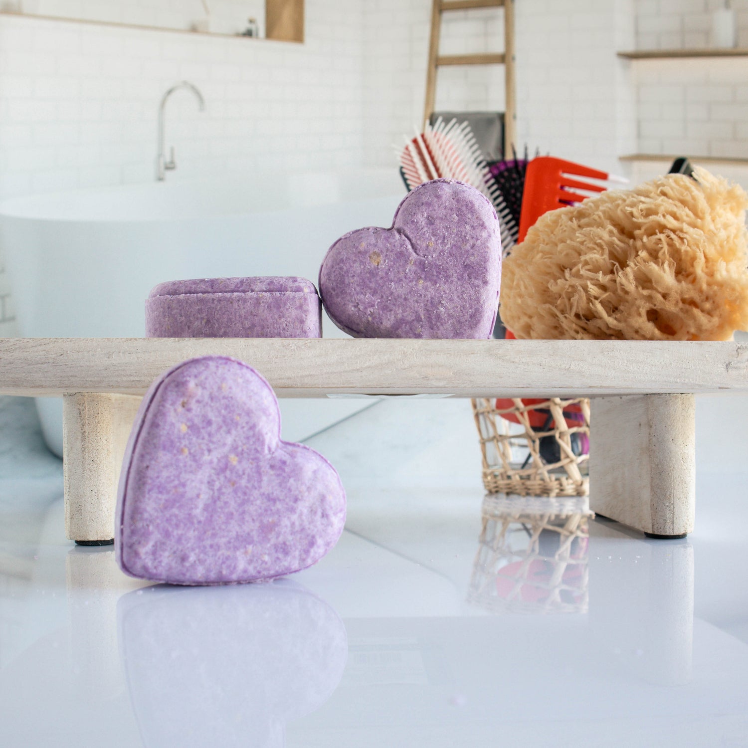 3 deep purple botanical bliss shampoo bars shaped in a heart. One is leaning on its side while the other two are on a trey, one leaning on the other. there is a loofah next to them with a cup of hair tools in the background
